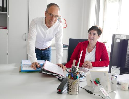 Ein Mann mit Hemd und eine Frau mit Blazer an einem Büroarbeitsplatz und blicken freundlich
